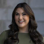 Sydnee Olsen, wearing a green top and with long wavy brown hair, smiles in front of a blurred gray background on the Bottarini Marketing Group site.